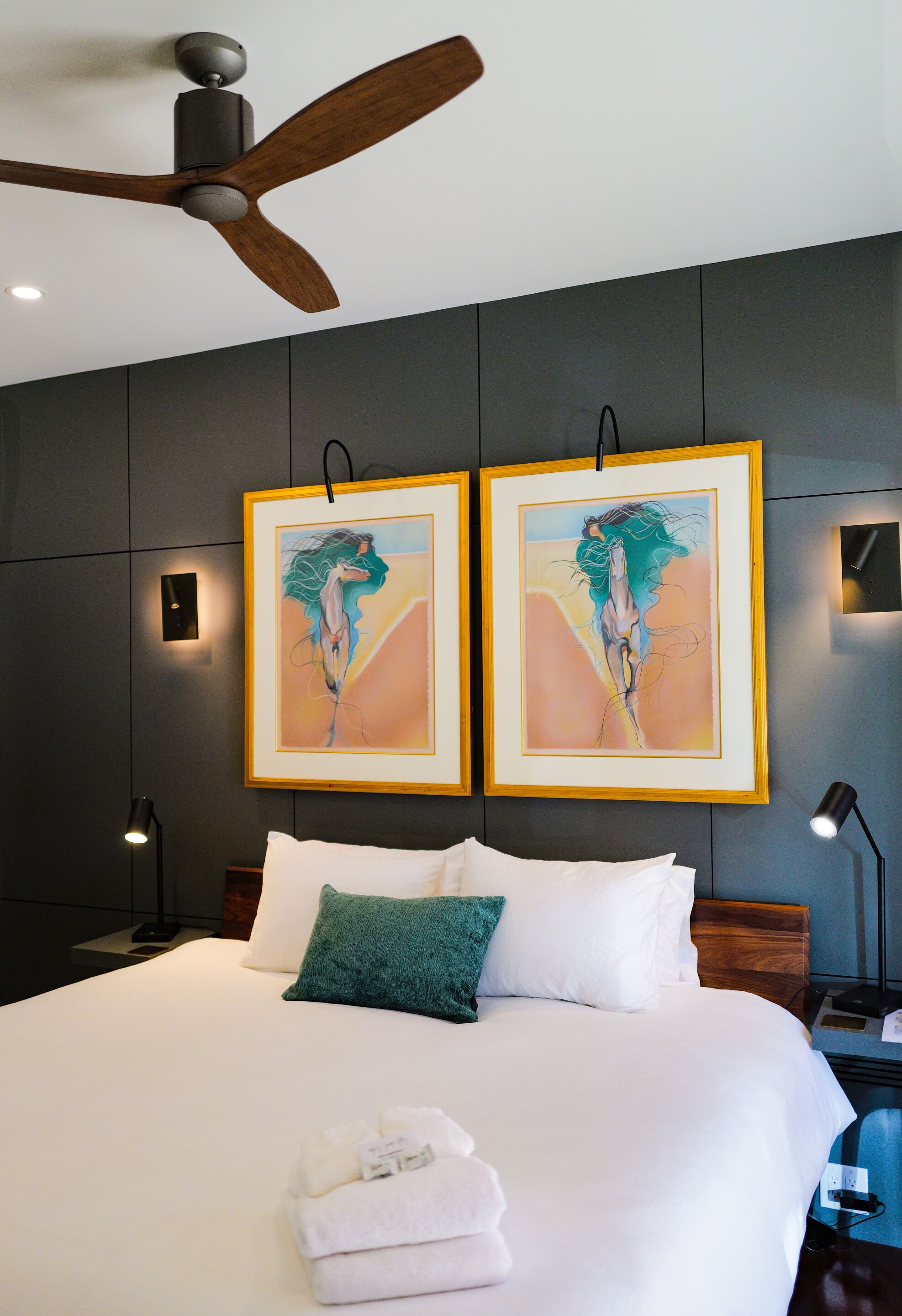 Een moderne hotelkamer met een groot bed, een plafondventilator, twee ingelijste kunstwerken, een nachtkastje en een bedlampje.