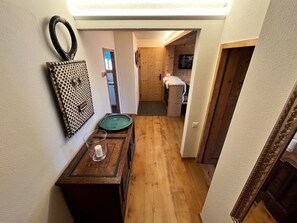 Ein schmaler Flur mit Holzboden, einem kleinen Tisch mit einem grünen Teller und einem Glas sowie einer Wanddekoration.