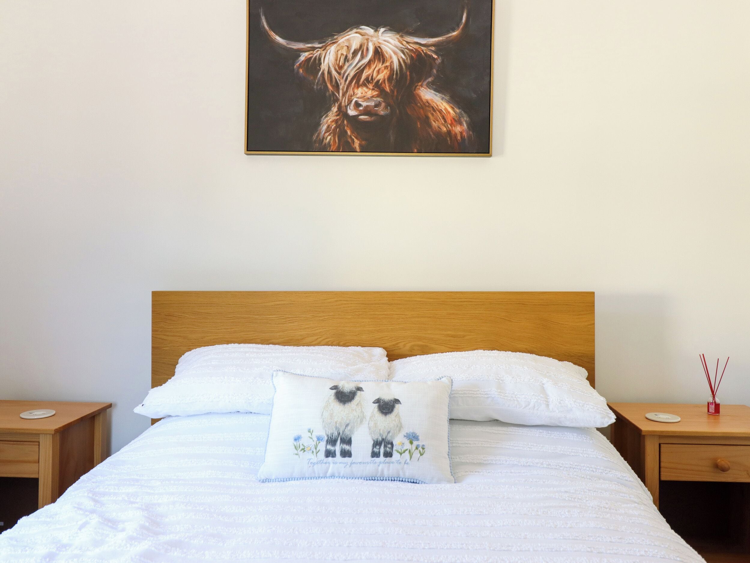 A bedroom with a wooden headboard, white bedding, two bedside tables, and a wall-mounted painting of a bull.