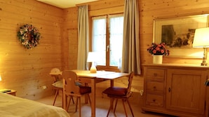 Ein gemütliches Zimmer mit Holzwänden, einem Bett, einem Esstisch mit Stühlen, einem Schminktisch und einem Fenster mit Vorhängen.