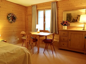 Ein gemütliches Zimmer mit Holzwänden, einem Bett, einem Esstisch mit Stühlen, einem Schminktisch und einem Fenster mit Vorhängen.