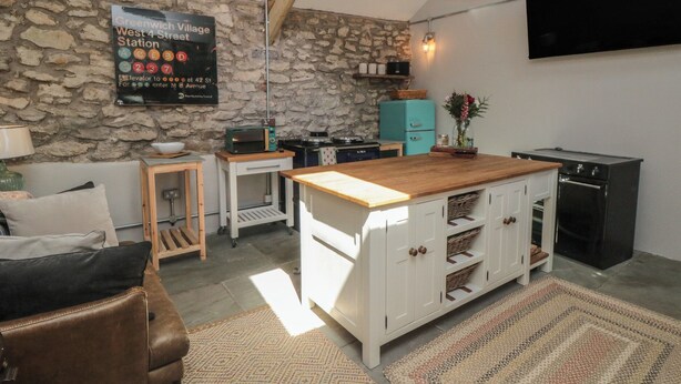 A kitchen with a stone wall, a wooden ceiling, a blue hanging light, a black TV, and a kitchen island with cabinets. - The Cottage at Thorpe Farm (York)