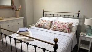 A neatly made bed with floral pillows, a bedside table with a lamp, and a dresser with a mirror and vase. - The Cottage at Thorpe Farm (York)