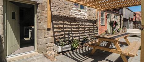 A stone building with a wooden pergola, a stone patio, and a wooden table.