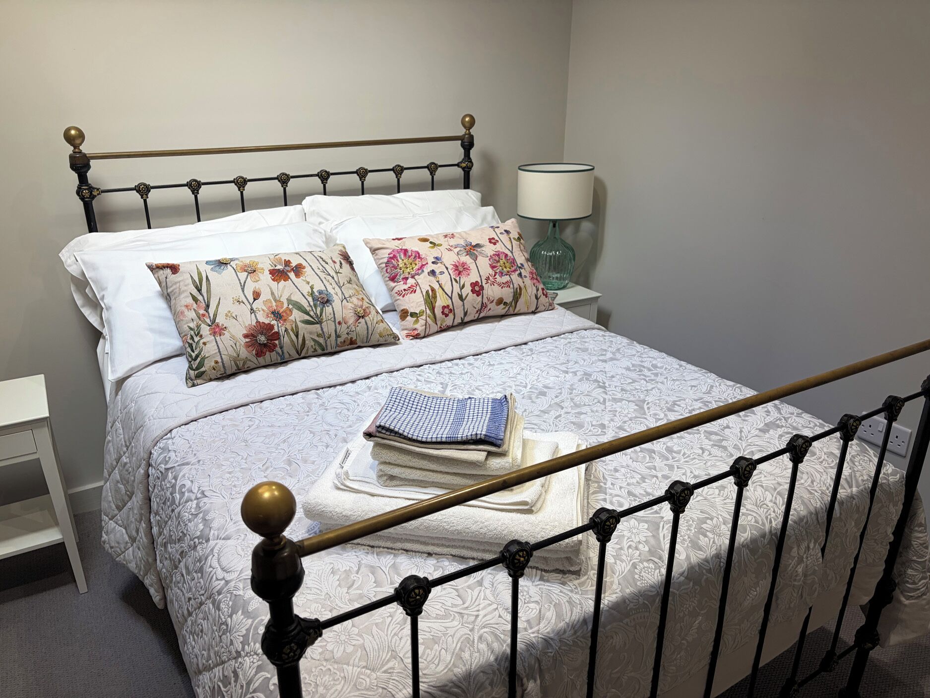 A neatly made bed with floral pillows, a lamp, and a bedside table with a folded towel.