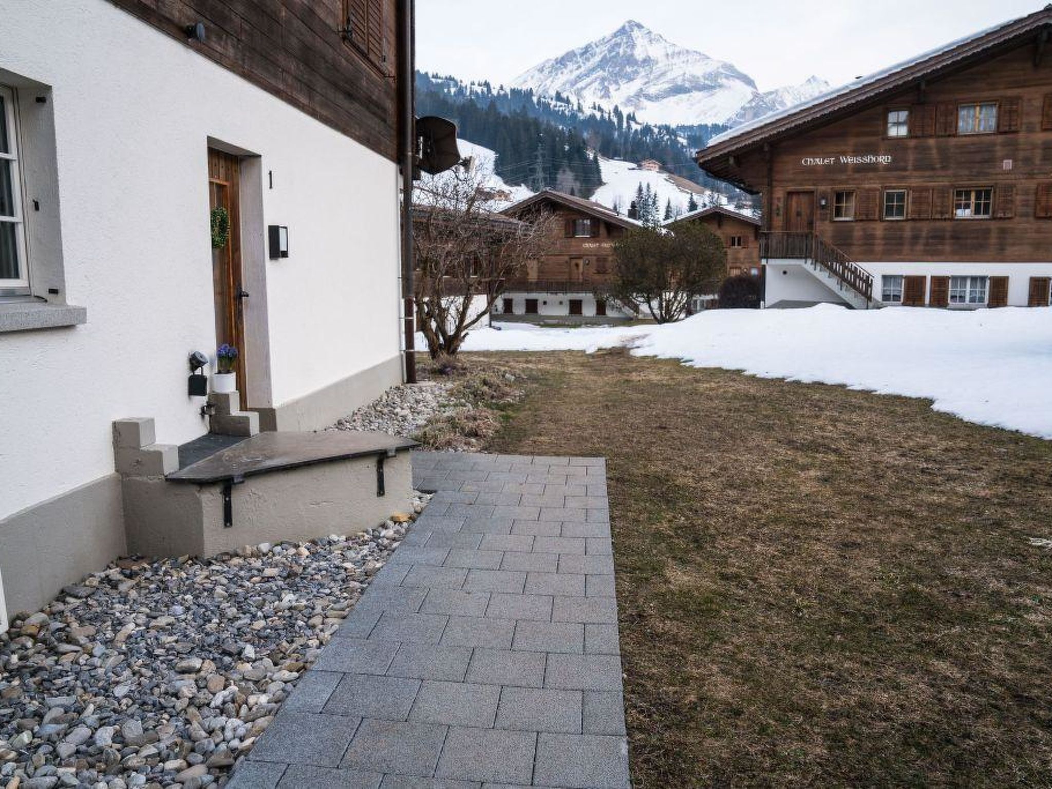A building with a stone pathway leading to a wooden door, surrounded by snow-covered ground and mountains in the background.