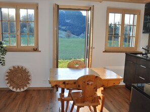 A kitchen with wooden cabinets, a dining table, and chairs. There are three windows with a view of a green landscape and mountains. - Tüllerhus Studio Studio, Dusche, WC 2-Bettwohnung by Interhome (Gstaad)
