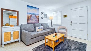 A living room with a sofa, a coffee table, a cabinet, a painting, a lamp, and a door. - Cozy Condo, Steps to St. Pete Beach + Shared Pool (St. Petersburg Beach)