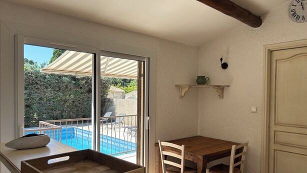 Eine Küche mit einem Holztisch, Stühlen, einem Holztablett, einer kleinen Ablage mit einem grünen Topf und Blick auf einen Swimmingpool.