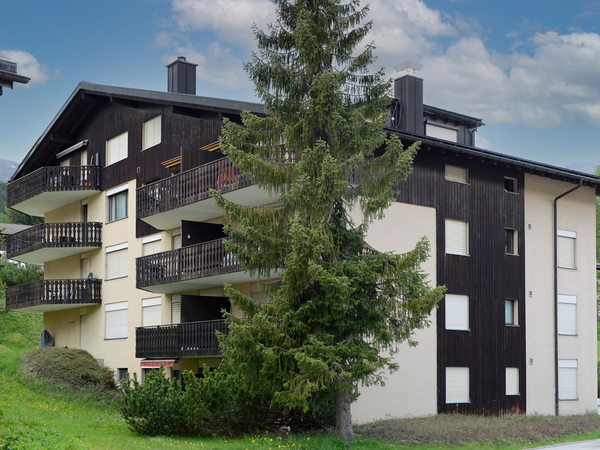 Un edificio de varios pisos con balcones, una chimenea y un gran árbol de hoja perenne al frente.