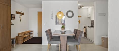 A modern dining area with a wooden table and grey upholstered chairs, a hanging light fixture, and a kitchen in the background.