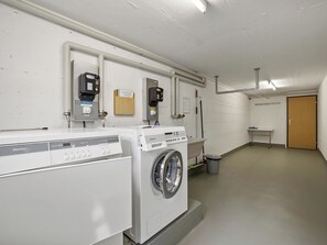 A laundry room with two front-loading washers and dryers, a trash can, and a small table with a chair. - A302 Appartement/Fewo Ferienwohnung A302 by Interhome (Lenzerheide)