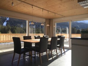 En matplats med ett träbord och stolar, stora fönster med utsikt över bergen och hängande lampor.