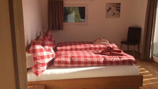 A bedroom with a wooden bed, checkered bedding, a window with a view, and a chair. - Chesa Parpan Appartement/Fewo Appartement/Fewo by Interhome (Valbella)
