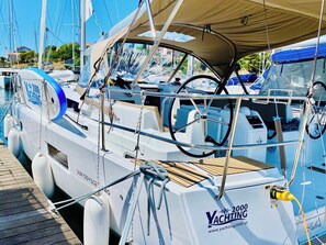 Eine Yacht mit dem Namen '2000 VYCTING' liegt in einer Marina festgemacht, wo sich mehrere Boote befinden. Man sieht ein Steuerrad und eine Webadresse.