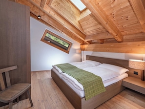 A bedroom with a wooden ceiling, a bed with white linens, a green throw, a nightstand with a lamp, and a window with a view of greenery.