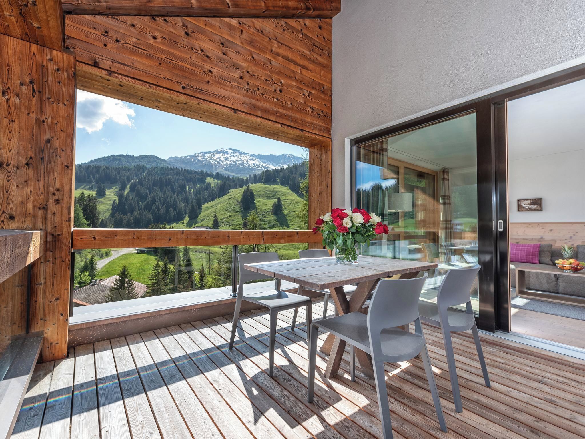 A wooden deck with a table and chairs, a vase of flowers, and a view of a mountainous landscape.