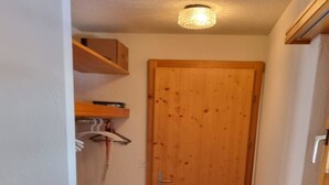 Ein schmaler Flur mit einer Holztür, einer schwarzen Matte im Eingangsbereich und einem Holzbodenregal mit einem weißen Handtuch.