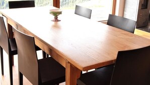 Mesa de jantar com seis cadeiras, uma tigela de frutas e vista para uma floresta através de grandes janelas.