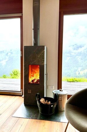 A wood-burning stove with a visible fire, a bucket of wood, and a view of a forested landscape through large windows. - Pas-Cheus Appartement/Fewo, Bad, WC, 2 Schlafräume by Interhome (Vaz)
