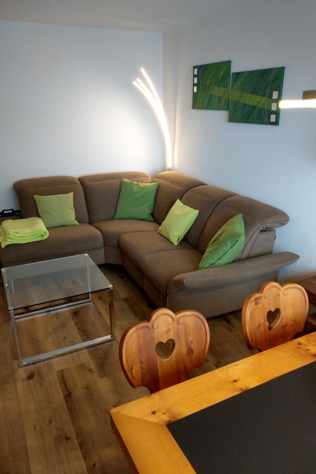 Ein modernes Wohnzimmer mit einem braunen Ecksofa, grünen Kissen, einem Glastisch und Holzstühlen mit herzförmigen Ausschnitten.