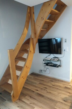 A wooden spiral staircase in a room with a flat-screen TV mounted on the wall and a telephone on a shelf below.