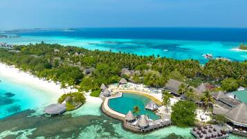 Vista aérea de un resort tropical con una piscina grande, bungalows sobre el agua y una playa.