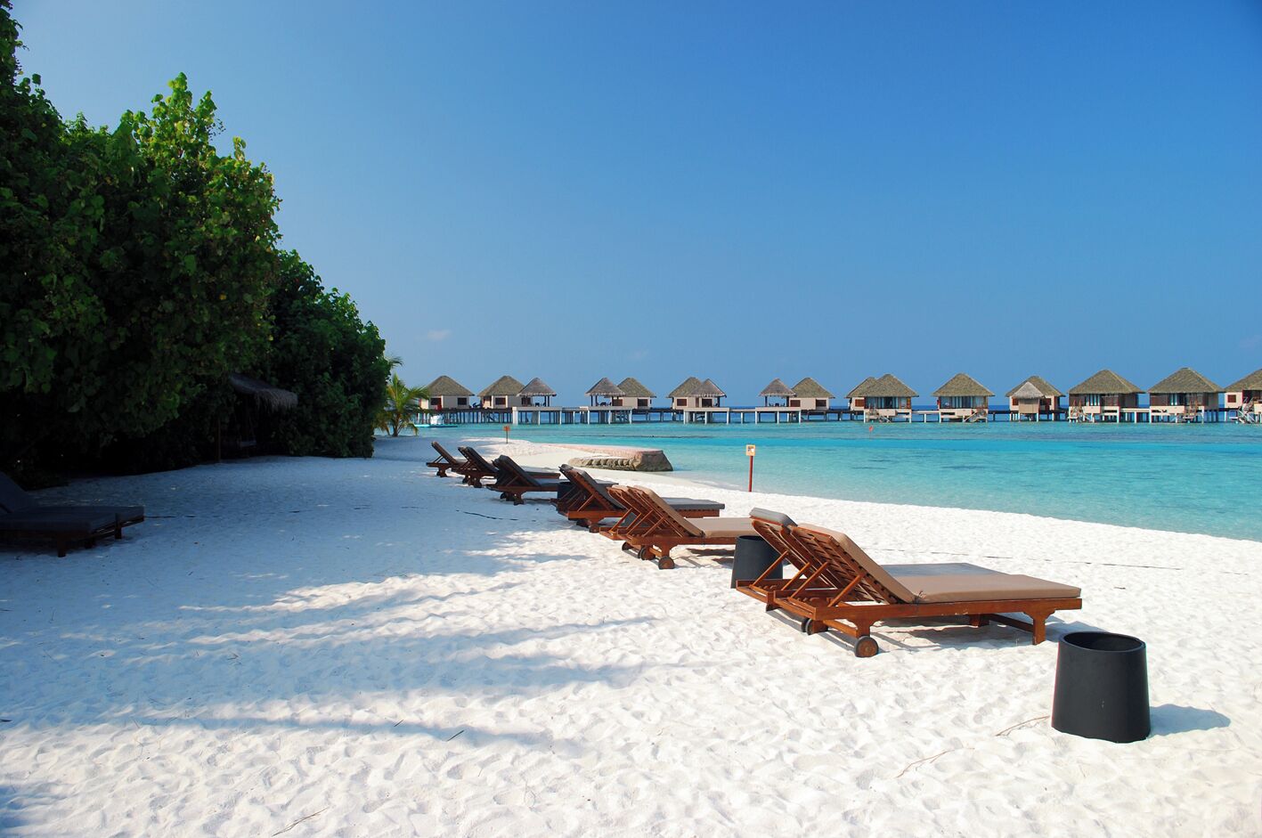 A beach with sun loungers, a trash bin, and overwater bungalows in the background.