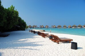 Ein Strand mit Liegestühlen, einem Mülleimer und Overwater-Bungalows im Hintergrund.