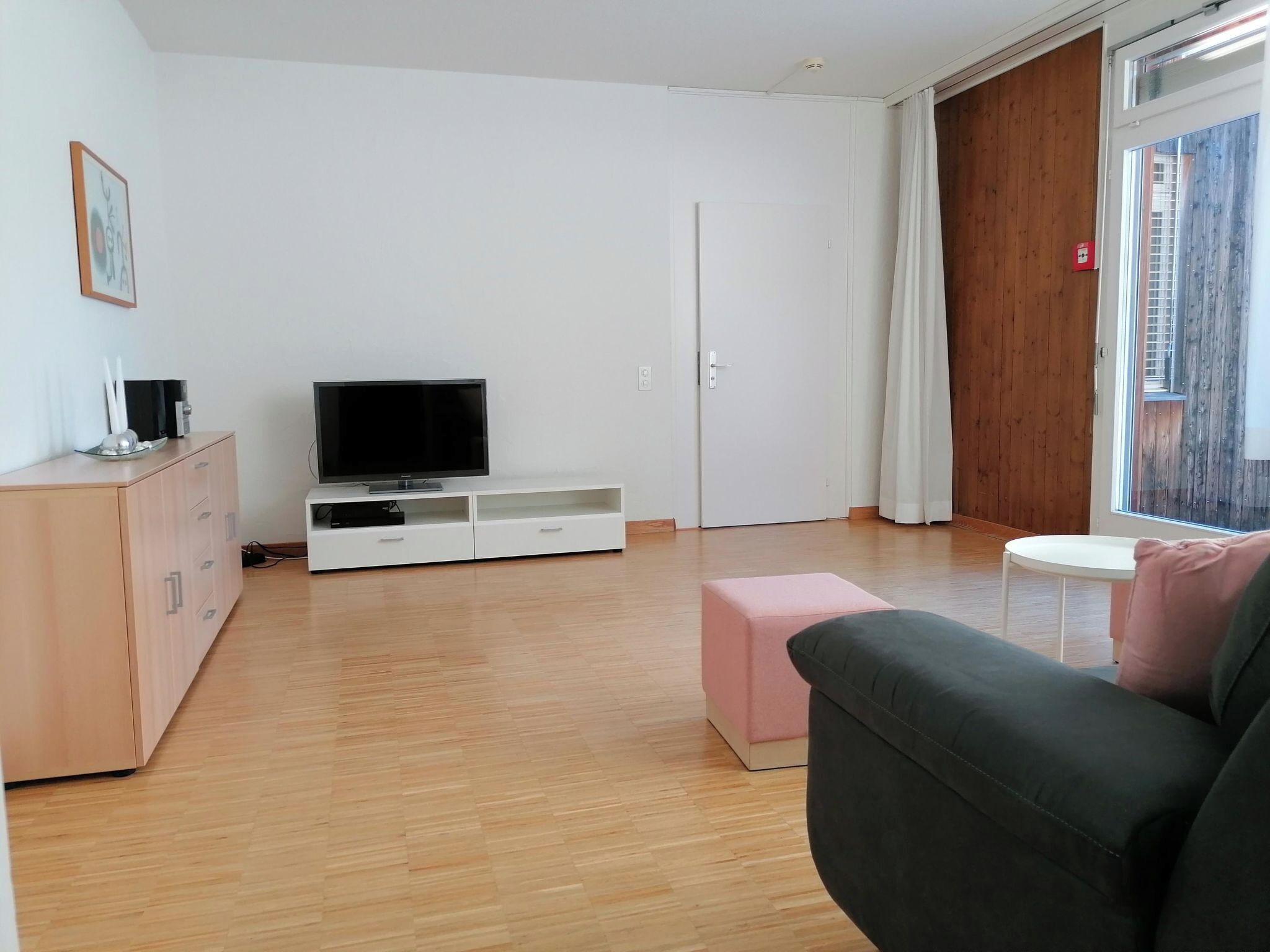 A modern living room with a flat-screen TV, a wooden cabinet, a pink ottoman, and a grey sofa.