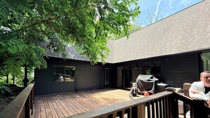 A wooden deck with a railing, a covered outdoor seating area, and a building with large windows and a dark exterior. - Private Mountain Spa Escape – Hot tub, sauna, Peloton & firelit lounges! (Wintergreen Resort)