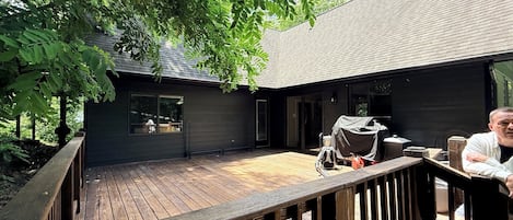 Una terraza de madera con barandilla, un espacio de estar al aire libre cubierto y un edificio con grandes ventanales y fachada oscura.