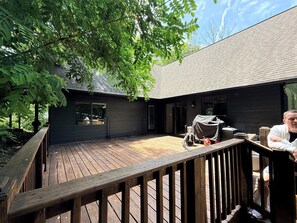 A wooden deck with a railing, a covered outdoor seating area, and a building with large windows and a dark exterior. - Private Mountain Spa Escape – Hot tub, sauna, Peloton & firelit lounges! (Wintergreen Resort)