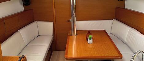A compact boat interior with a wooden table, white cushioned seating, a small potted plant, a mounted TV, and a glass sink.