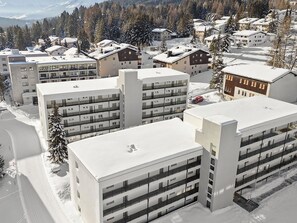 A modern multi-story building with a flat roof, likely a hotel or apartment complex, surrounded by snow-covered trees and a mountainous backdrop. - Nr.43 Haus Sotmirana Appartement/Fewo Nr.43 Haus Sotmirana by Interhome (Lenzerheide)