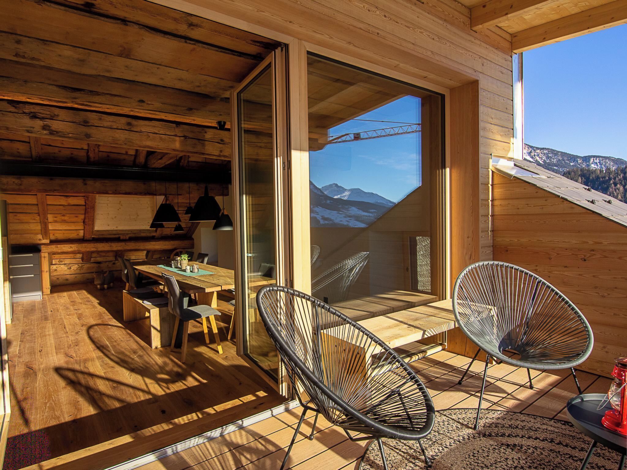 Une cabane en bois avec un coin repas, deux chaises en osier et une vue sur les montagnes, visible à travers une porte vitrée.