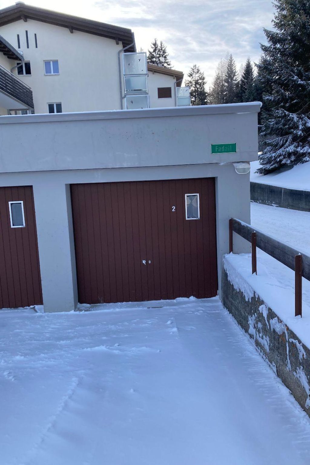 Un edificio con dos puertas de garaje, suelo cubierto de nieve y una valla de madera.