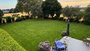 Um gramado bem cuidado com um espaço de terraço com mesa e cadeiras, rodeado por diversos arbustos e árvores.