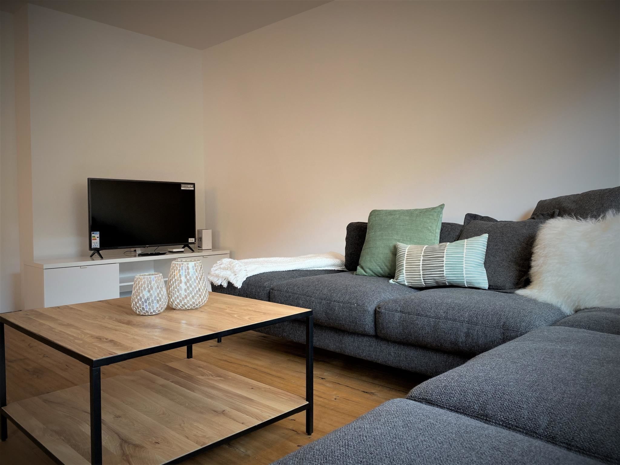 Una sala moderna con un sofá gris, un televisor de pantalla plana, una mesa de centro de madera y jarrones decorativos.