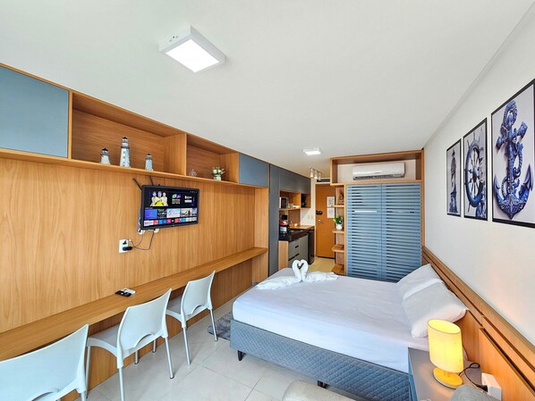 A hotel room with a bed, bedside table, lamp, TV, desk, chairs, wardrobe, and artwork.