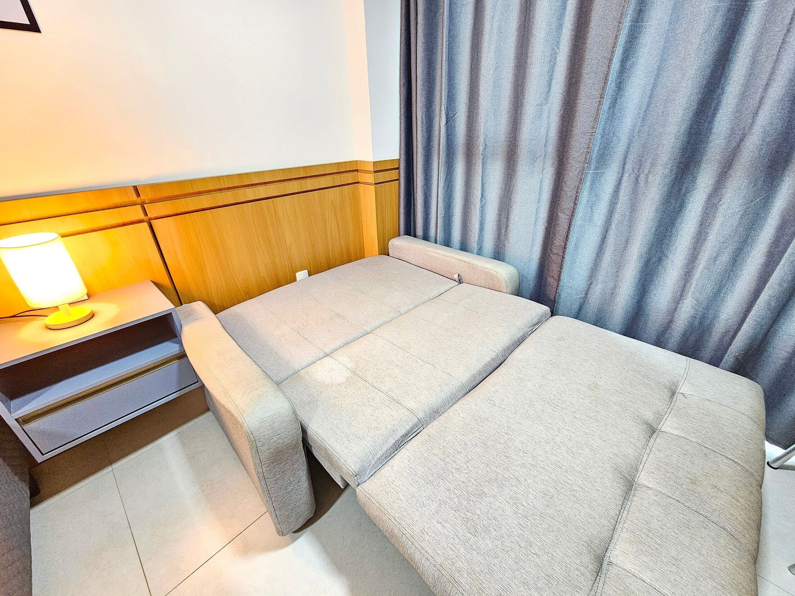 Quarto compacto com cama, mesa de cabeceira e luminária, além de cortina.