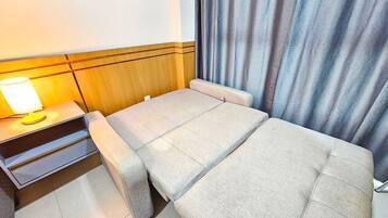 Quarto compacto com cama, mesa de cabeceira e luminária, além de cortina.