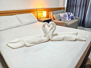A hotel room with a neatly made bed, a nightstand with a lamp, a sofa with decorative pillows, and a window with curtains.