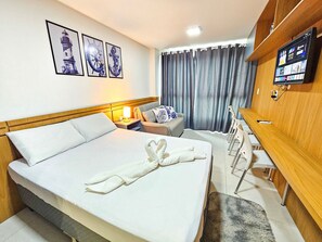 A hotel room with a bed, a desk, a chair, a TV, and artwork on the wall.