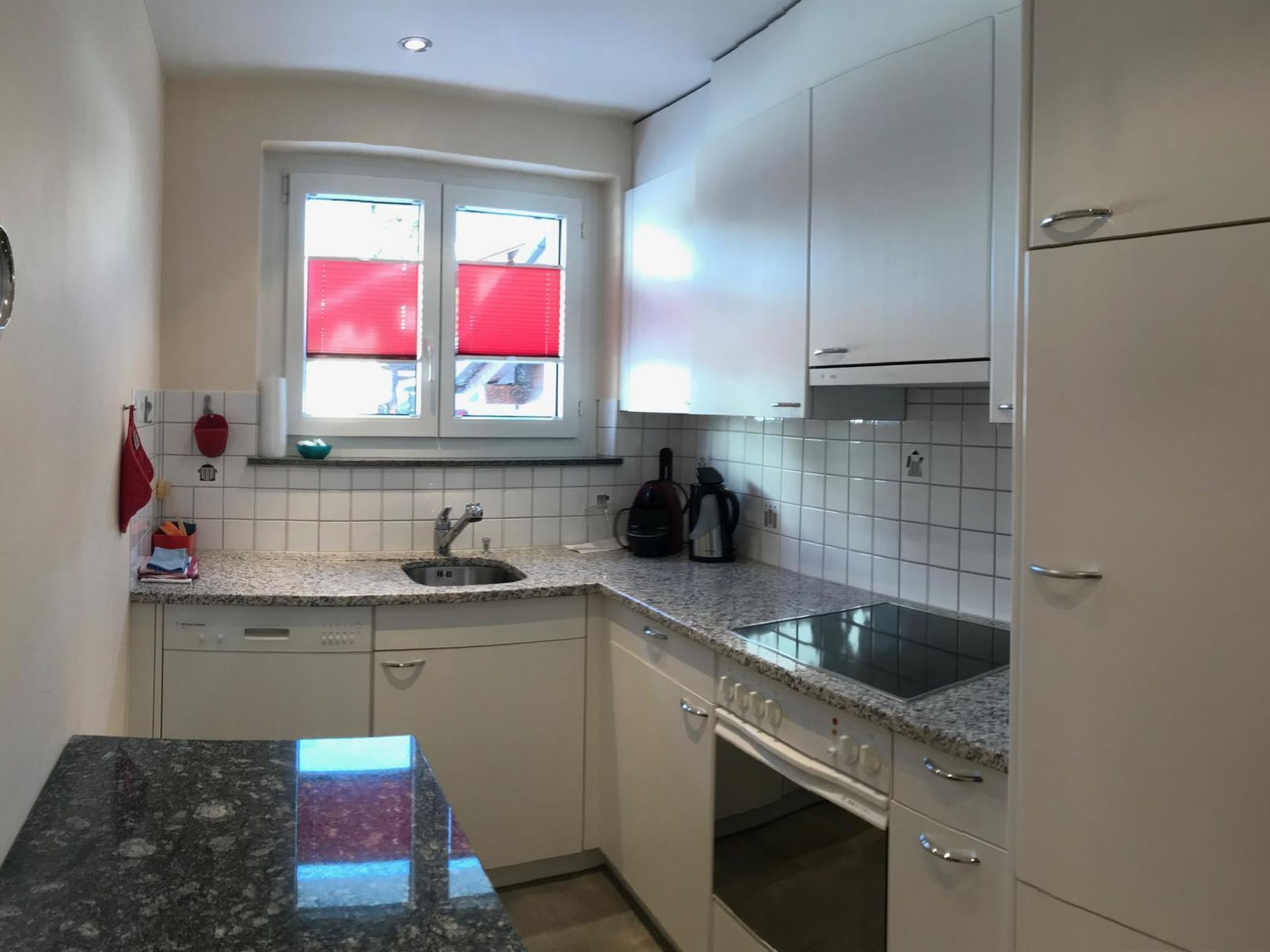 A compact kitchen with white cabinets, a granite countertop, a built-in oven, and a window with red blinds.