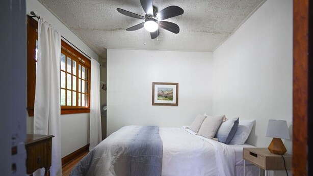 Um quarto com cama, ventoinha no teto, mesa de cabeceira com abajur e janela com cortinas.