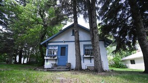 Uma pequena casa térrea com porta e janelas azuis, cercada por árvores e grama.