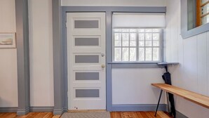 Um quarto com porta branca, janela com persianas, um banco de madeira e um tapete na porta.