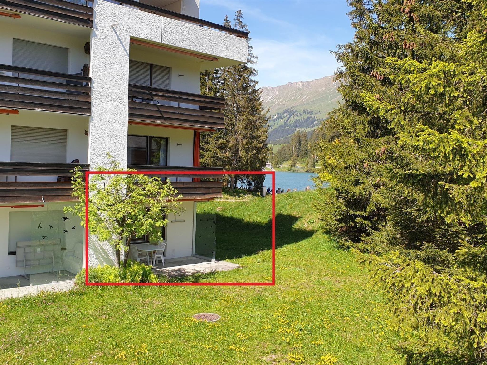 Ein modernes Gebäude mit Balkon, einer kleinen Sitzgelegenheit im Freien und Blick auf einen See und Berge.