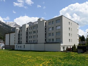 A multi-story building with a flat roof, large windows, and a grassy area with yellow flowers in front. - Nr.14 Haus Astoria Appartement/Fewo Nr.14 Haus Astoria by Interhome (Lenzerheide)